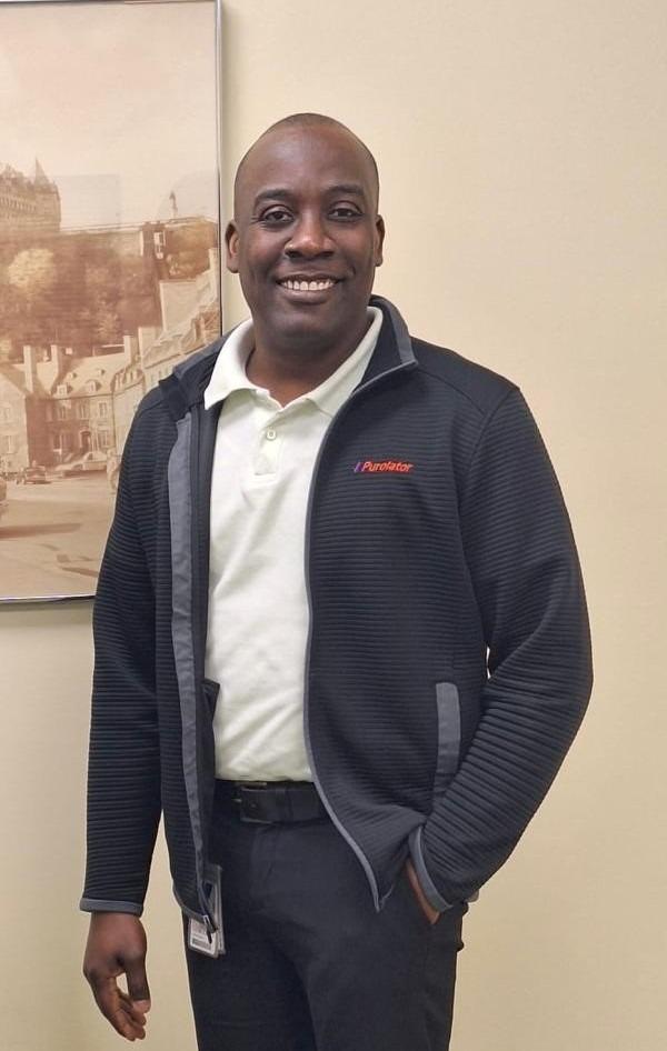 Walner Charles posing in front of a beige wall with a half of a framed photo hanging off to the left side.